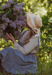 Vintage. Beautiful young woman in retro clothes, straw hat and with a bouquet of lilacs in a blooming garden. Boho style. Visual for content.