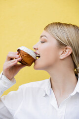 young woman eating cupcake with white glaze isolated on yellow.