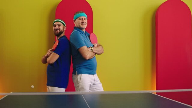 In front of the camera after the table tennis game posing happy and excited two plainer young and old men they holding the racket and looking straight