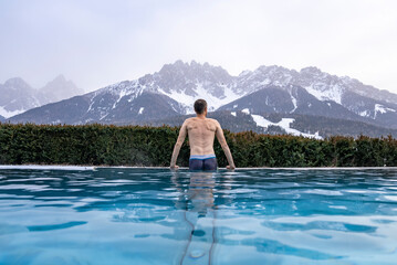 Tourist swimming in hot pool with view of mountains. Rear view of man enjoying in water at luxurious resort. Snow covered landscape against sky during winter.