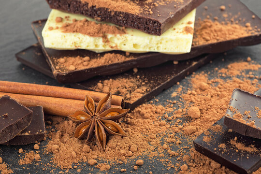 Broken chocolate pieces, cocoa powder, cinnamon sticks and anise stars on black stone background