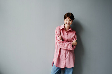Portrait of happy young Caucasian woman in oversize pink shirt standing with crossed arms against gray wall
