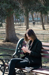 Fototapeta na wymiar A beautiful happy girl uses a mobile phone while sitting on a park bench. Vertical photo.