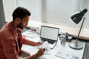 Serious young mixed race project manager with beard sitting at desk with laptop and examining charts in office