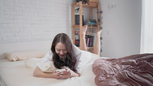 A young beautiful smiling woman is lying on the couch in the morning and typing a message on the phone. Lifestyle