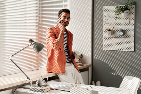 Portrait Of Confident Handsome Young Black Man In Stylish Outfit Standing With Takeout Coffee At Window And Chatting By Phone In Office