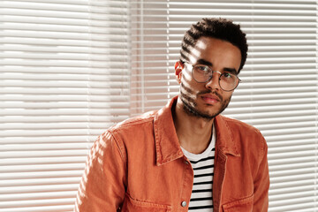 Fototapeta premium Portrait of serious young mixed race guy in round glasses and coral shirt standing against closed blinds