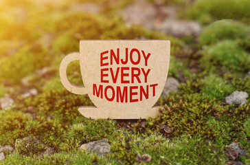 On the moss in the rays of the sun, a cup with the inscription - ENJOY EVERY MOMENT