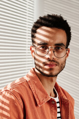 Portrait of serious young black man in round-shaped eyeglasses and coral shirt sitting against closed blinds