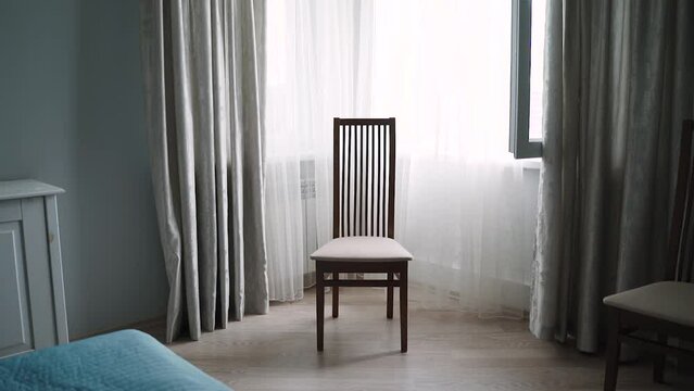 Wooden chair with slatted back stands in corner of cozy room near window with curtains. Day, window sash is open, wind shakes window tulle in bedroom. 