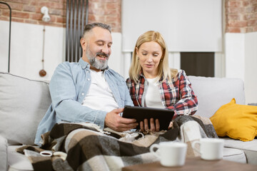 Mature people in love dressed in casual attire resting together on comfy couch with digital tablet in hands. Caucasian family surfing internet during leisure time at modern apartment.