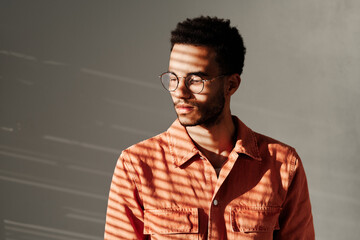 Calm young mixed race man in round-shaped glasses standing under light through blinds, isolated background