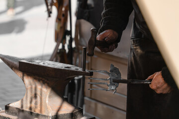 A blacksmith in a black leather apron holds a metal billet on an anvil in one hand and a hammer in...