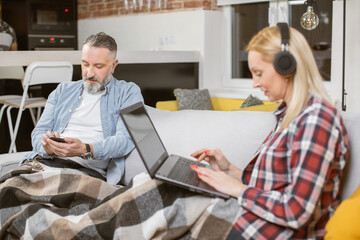 Caucasian middle aged man talking on smartphone while sitting on couch. Beautiful woman in headphones typing on modern laptop. Family relaxing at home with gadgets.