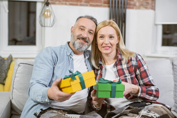 Portrait of charming mature woman and handsome man sitting closely on couch with gift boxes in hands and looking at camera. Happy family celebrating anniversary at home.