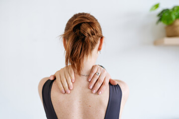 A woman with red hair stands with her back to the camera and massages her shoulder with two hands