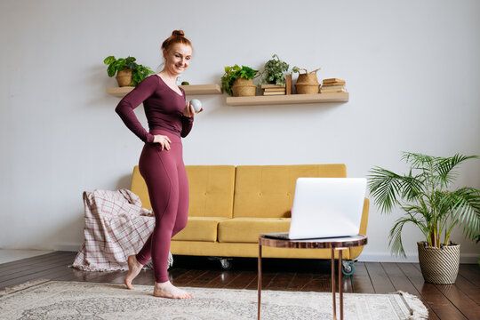 a red-haired woman leads an online pilates class in front of a laptop at home