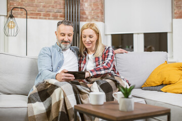 Positive mature couple in casual wear resting together on cozy sofa and using modern smartphone. Caucasian family of two surfing internet during free time at home.