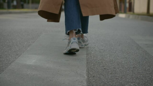 Rare view with low angle on feet of young beautiful fancy, stylish and confident female walking down the street wearing casual beige trench coat, blue jeans, white sneakers in city street on weekend