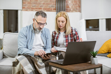 Obraz premium Serious caucasian couple planning domestic finances using modern laptop and smartphone. Mature man and woman in casual clothes sitting on couch with papers around.