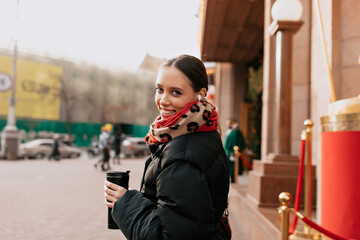 Fototapeta premium Wonderful smiling girl with dark hair in black jacket is walking on city in sunshine with coffee