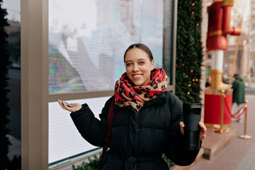 Charming smiling happy girl in warm dark jacket is smiling at camera and waving hands while walking around the city center 