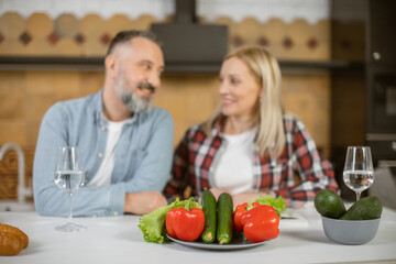 Blur background of caucasian married couple smiling to each other while standing together on domestic kitchen. Focus on table with healthy eco vegetables on plate.