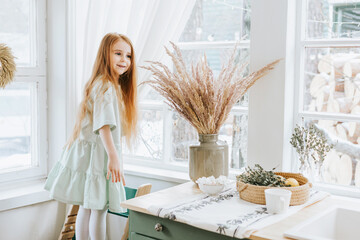 little red haired girl looks out the window in kitchen of rustic country house with beautiful...