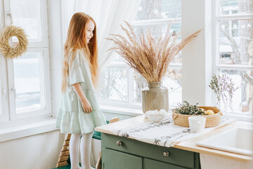 little red haired girl looks out the window in kitchen of rustic country house with beautiful...