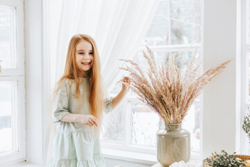 little red haired girl looks out the window in kitchen of rustic country house with beautiful...