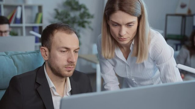 Young blond attractive woman in formal wear approaching colleague's desk to discuss presentation. Unshaven good-looking man in black suit listening to ideas. Office workers in modern office. Business
