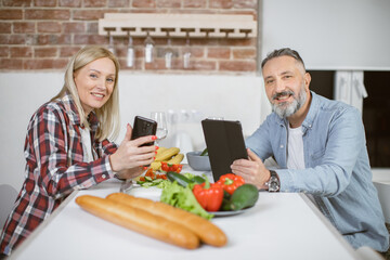Happy mature married couple smiling and looking at camera while sitting on kitchen with cell phone and tablet in hands. Caucasian family at home using modern gadgets.
