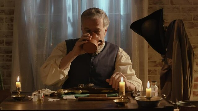 A mature bearded man from the 18th century takes a sip of coffee before he starts the process of cleaning his flintlock pistol.