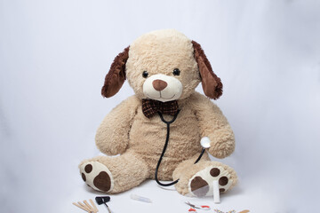 teddy bear with a beige bow tie with a stethoscope and medical items on a greyish white background.