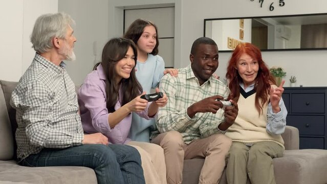 A large multi-ethnic family of different age generations play a video game at home sitting on the couch. Game On, Family Meeting, Multi Ethnic Family, Different Generations.