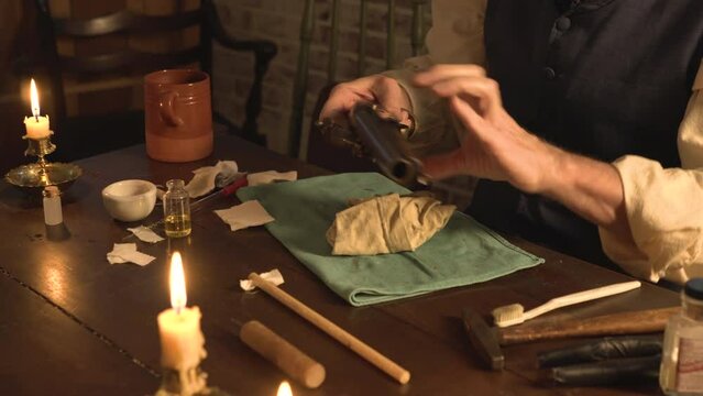 Lit by candlelight and firelight a man from the 1700s polishing his flintlock pistol as part of maintenance on the firearm.