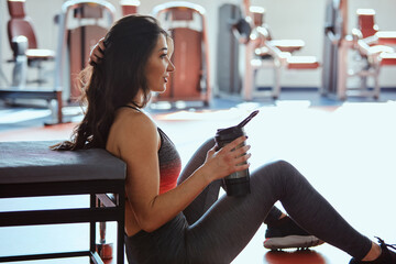 A sportswoman sits on the floor in a gym and drinks a protein shake for strength. - obrazy, fototapety, plakaty