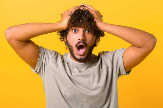 Excited shocked Indian man holding hands on head and screaming looking at camera with big eyes and open mouth, shocked amused with news, win at lottery. Indoor studio shot isolated on yellow