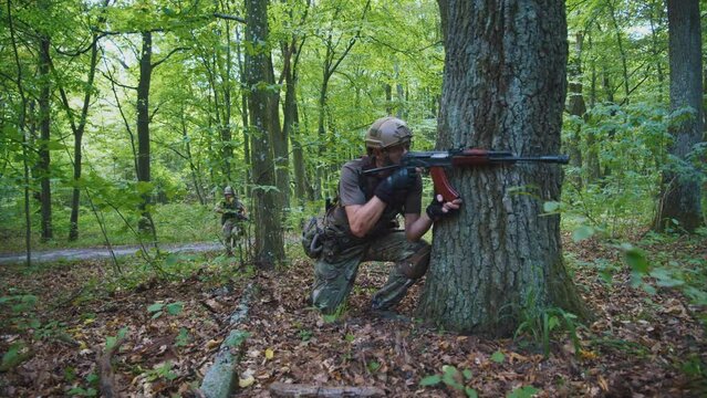 Close up soldier man in camouflage shoots machine gun sitting near tree in forest. Military concept. On background running soldiers