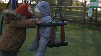 Obraz premium Funny Little Preschool Chubby Girl Older Sister Rides Younger Newborn Infant Brother Boy On Swing. Baby Playing On Playground. Kids Entertainment, Childhood, Child Development, Happy Family Concept