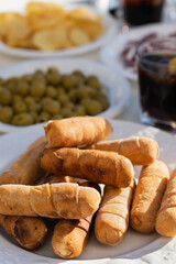Table full of snacks in a sunny day with some drinks. Tequeños in the front with other aperitivos, like olives and crisps. Drinking vermouth. 