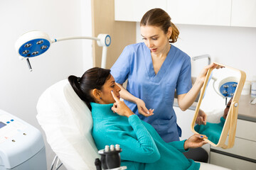 Obraz premium Hispanic woman holding mirror and pointing finger on her face during cosmetologist's examination in clinic.