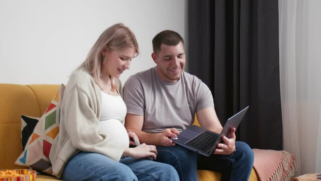 A happy couple is sitting on the couch and talking via video link from a laptop and showing a photo of an ultrasound of pregnancy. A young woman is pregnant. Young family