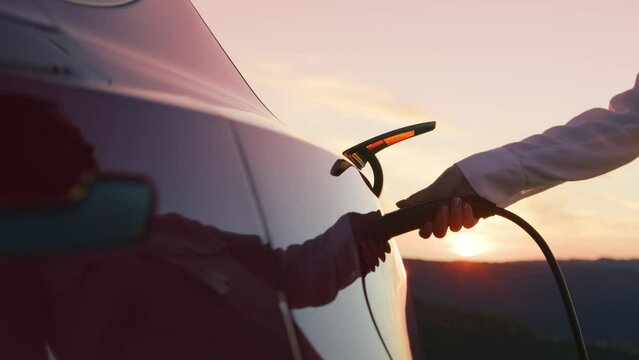 Slow motion woman plugging in power cable in 0 emission electric car at charging station with nature landscape at pink sunset. Cinematic 4K environmental eco-friendly technologies, climate change shot