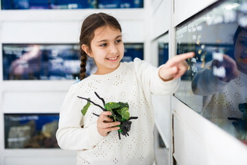 Portrait of interested cute girl pointing with finger on aquarium with tropical fishes in pet shop