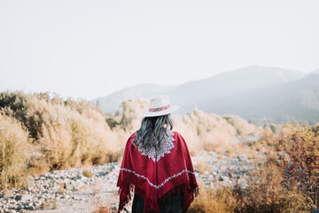 Traditional latin american woman wearing a red poncho and a hat seen from behind, in a natural...