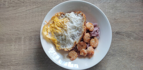 Garlic Fried Prawns and Fried Egg with Rice, placed in a white plate, on a wooden table. From above: Purple rice comes from well brown rice.