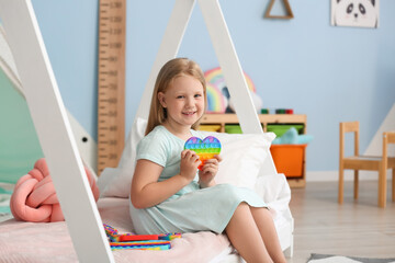 Happy little girl sitting on bed and holding heart shaped pop it fidget toy in child room - obrazy, fototapety, plakaty