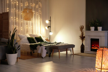 Interior of stylish bedroom with modern lamp, houseplants and fireplace in evening