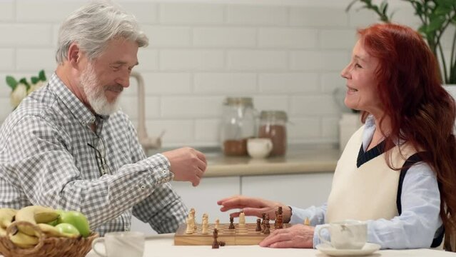 Happy Senior Couple 60-70 Years Old Playing Chess At Home Sitting At The Table In The Kitchen. Game On, Family Meeting, Multi Ethnic Family, Different Generations.